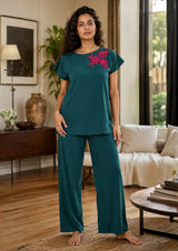 Woman wearing teal pajama set with floral embroidery in a living room.